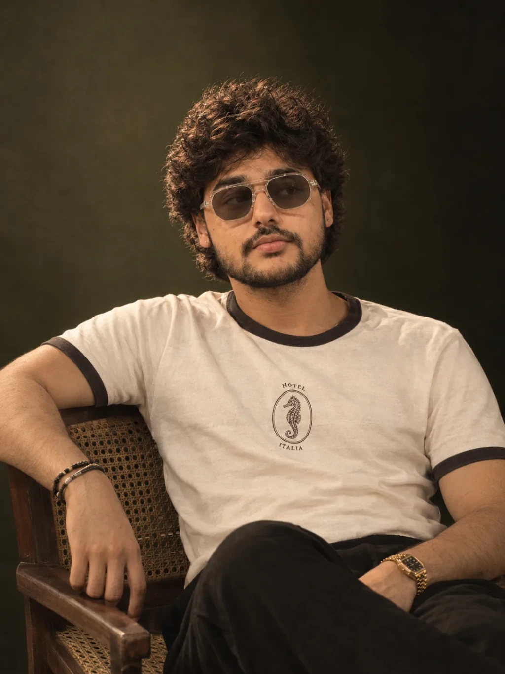 A refined, warm studio portrait with an olive backdrop, relaxed chair pose.